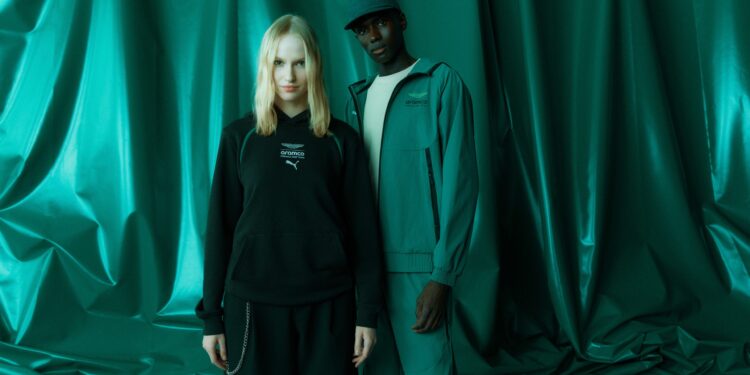 Two people stand in front of a green draped background; one wears a black hoodie and skirt, while the other sports a green jacket, pants, and cap from the PUMA Motorsport F1 Team Collection.