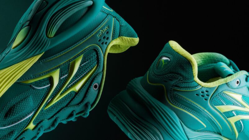 Two teal and lime green athletic sneakers from the PUMA Motorsport F1 Team Collection are shown against a dark background, highlighting their textured fabric, intricate design, and thick soles.