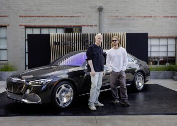 Two people stand in front of a black Mercedes-Maybach parked on a platform with a modern backdrop in an outdoor setting, resembling a rolling music studio.