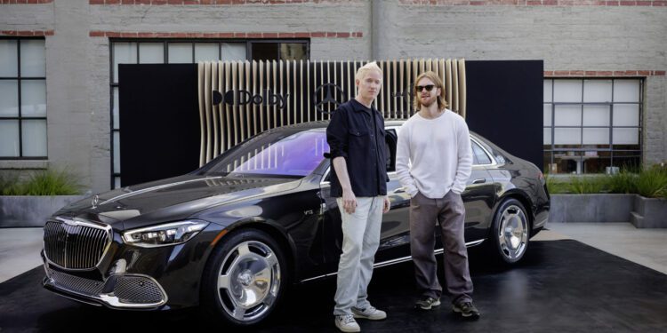 Two people stand in front of a black Mercedes-Maybach parked on a platform with a modern backdrop in an outdoor setting, resembling a rolling music studio.