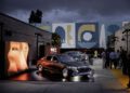 A luxury Maybach is displayed on a lit outdoor platform at an evening event, with people gathered nearby and abstract murals on the wall in the background.