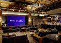 Modern lounge with large digital screens displaying Mercedes, Maybach, and Universal logos, featuring cozy seating, wooden decor, and a person working behind a sleek bar counter, evoking the vibe of an upscale music studio.