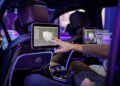 A person interacts with a touch screen displaying a 3D interface in the backseat of a Mercedes, with another screen visible in the background.