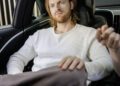 A man with long hair and a beard sits in the backseat of a Mercedes, looking forward with a neutral expression. Another person's hand is partially visible in the foreground.