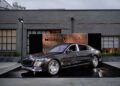 A sleek black Mercedes luxury sedan is displayed on a platform outdoors, in front of a modern industrial-style building that could double as a chic music studio.