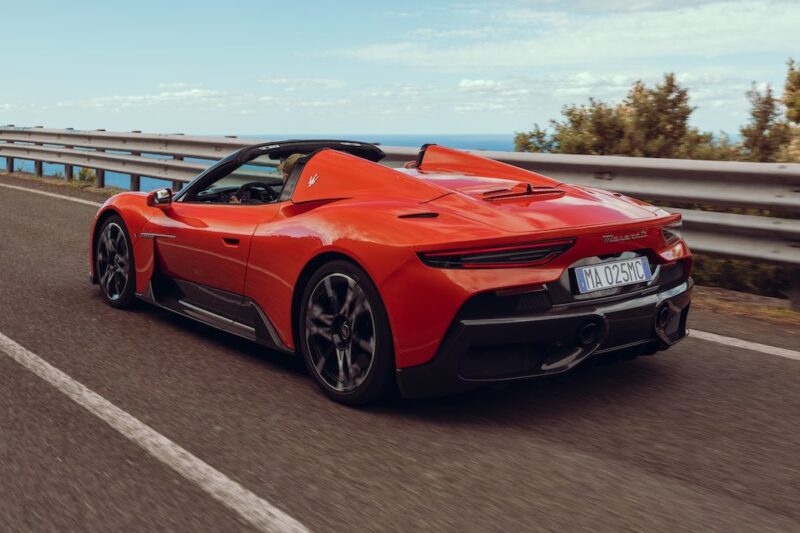 A 2026 Maserati MCPura Cielo First Drive shows a red sports car gliding down a paved road beside a metal guardrail, framed by trees and a blue sky in the background.