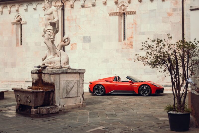 A 2026 Maserati MCPura Cielo First Drive red sports car is parked near a stone fountain and statue in a paved courtyard with a marble wall in the background.