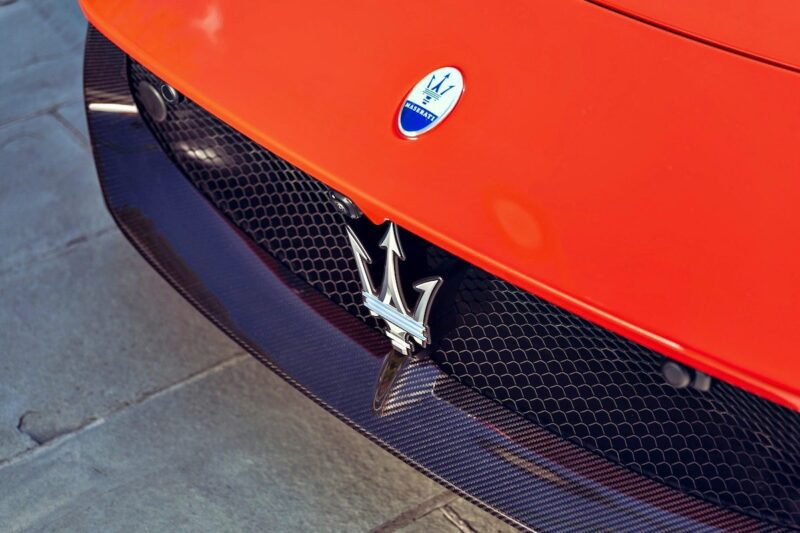 Close-up of the front grille of an orange 2026 Maserati MCPura Cielo First Drive, featuring the iconic Maserati trident emblem and badge set against a striking mesh grille background.