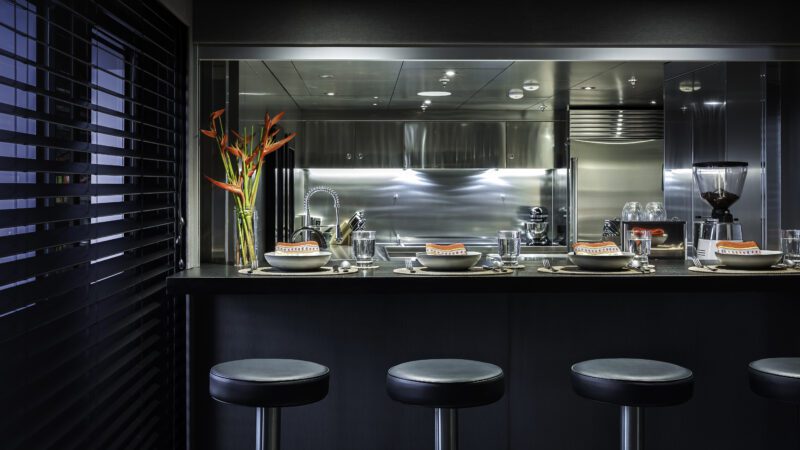 Modern kitchen with a sleek black breakfast bar, four stools, place settings for four, stainless steel appliances, and a vase with red flowers—stylish enough to grace an IKONIK Yachts power catamaran like THE BEAST.