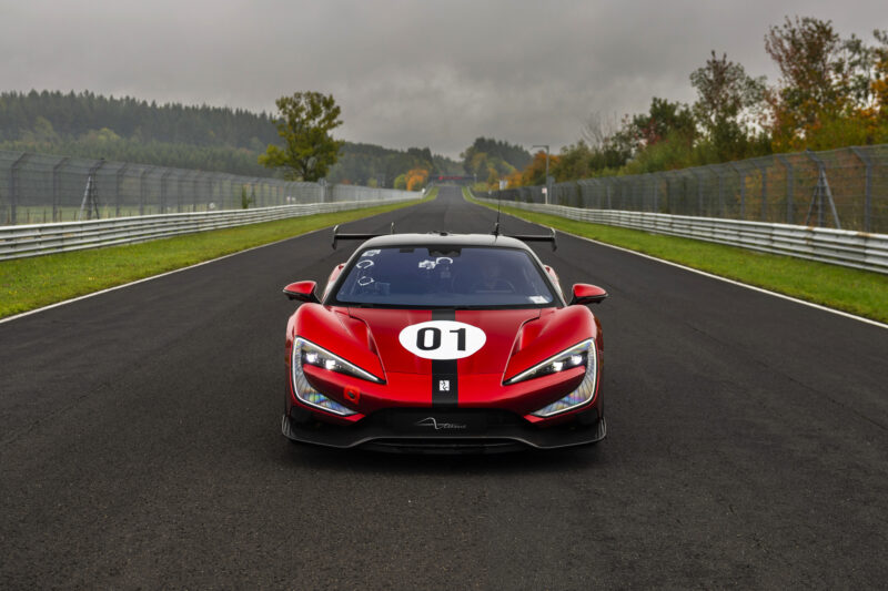 A red sports car with the number 01 on its hood, reminiscent of electric hypercars, is parked on a racetrack, surrounded by trees and a cloudy sky.