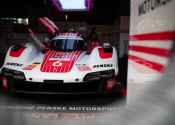 A Porsche Penske Motorsport race car with red and white livery is parked in a garage, with the driver's door open and team branding visible.