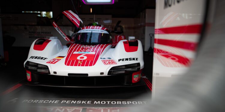 A Porsche Penske Motorsport race car with red and white livery is parked in a garage, with the driver's door open and team branding visible.