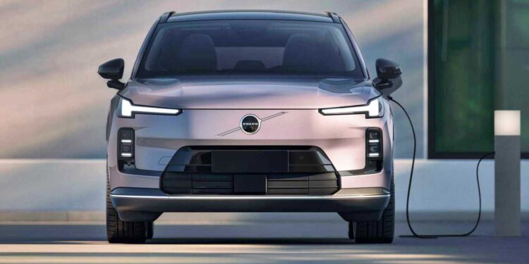 Front view of a light pink Volvo electric SUV parked and charging at a station next to a modern building, reflecting Volvo’s global plug-in hybrid sales crossing one million.