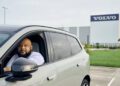 A man sits in the driver's seat of a parked SUV in front of a building with a large Volvo sign on it.
