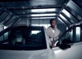 A woman wearing a gray coat by Sergio Hudson stands beside an open Volvo EX90 car door inside a modern, metallic garage.