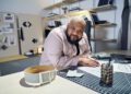 A person leans over a worktable covered with sketches, fabric, and a belt in Sergio Hudson’s well-organized design studio, looking directly at the camera.