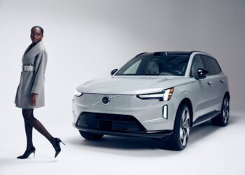 A woman in a Sergio Hudson grey coat and heels stands next to a silver Volvo EX90 SUV in a studio with a plain light background.