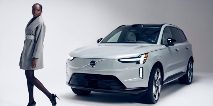 A woman in a Sergio Hudson grey coat and heels stands next to a silver Volvo EX90 SUV in a studio with a plain light background.