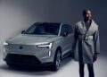 A person in a gray coat stands in front of a silver Volvo EX90 SUV in a studio setting with neutral lighting, embodying the refined elegance of Sergio Hudson’s style.