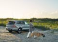 A person walks a large dog near a silver Volvo EX90 parked on a sandy path with grassy dunes in the background at sunset.