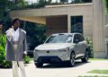 A person stands outside near a modern house, where a silver Volvo EX90 SUV by Sergio Hudson is parked in the driveway and connected to an electric vehicle charging station.