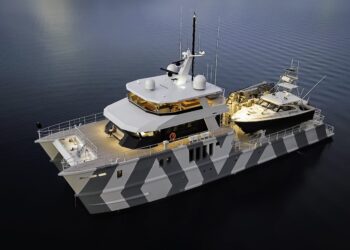 A large, modern power catamaran from IKONIK Yachts with a camouflage-patterned hull is anchored in calm water, carrying a smaller boat on its deck—an impressive vessel aptly named THE BEAST.