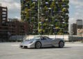 A silver Pagani Huayra Codalunga is parked on a paved area with modern buildings and a vertical garden in the background under a partly cloudy sky, capturing the spirit of the Las Vegas Concours.