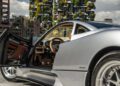 A silver Pagani Huayra Codalunga with its driver-side door open reveals a tan and black interior, parked in an urban area with modern buildings and greenery, reminiscent of a Speedster showcased at the Las Vegas Concours.