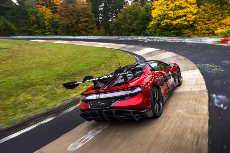 A red Electric Hypercar with a large rear wing drives on a curving racetrack surrounded by trees with green and autumn-colored leaves.