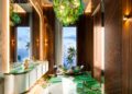 Luxurious bathroom at Aureum Estate with green marble floor, modern white vanity, sculptural bathtub, large windows showing palm trees and ocean, and a green glass chandelier overhead—true Dubai Hills elegance.