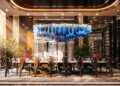 A modern, luxurious dining room at Aureum Estate features a long table with dark chairs, a blue geometric chandelier, lush plants, and decorative lighting accents inspired by the opulence of Dubai Hills.