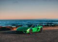 A green Lamborghini Centro Stile at 20 sports car is parked on a coastal road near the ocean, with rocky shorelines and distant hills under a clear sky.