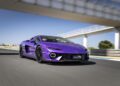 A purple Lamborghini speeds along a racetrack under a bridge, its vibrant colors blurred with motion beneath a clear blue sky.