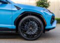 Close-up of the front left wheel and fender of a blue Lamborghini Urus SUV, showcasing the black alloy rim, tire, and part of the headlight, highlighting the bold colors and striking design origins of Lamborghini.