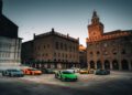 A group of colorful sports cars, including a striking Lamborghini, is parked in a historic European city square with old buildings and a clock tower at dusk.