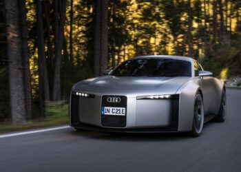 A silver Audi Concept C, a street-legal production sports EV, drives on a forested road during daylight, showcasing its modern, minimalist design and illuminated headlights.