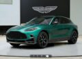 An updated green Aston Martin SUV is displayed on a circular platform in a modern showroom with the Aston Martin logo behind, offering inspiration for your own design using the online configurator.