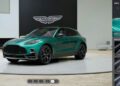 A green Aston Martin DBX SUV is displayed on a circular platform indoors, with customization options from the online configurator visible on the right and the Aston Martin logo in the background.