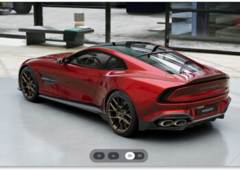 A red Aston Martin sports car with bronze alloy wheels is parked indoors on a concrete floor near large windows, capturing the refined look you can create with the Aston Martin online configurator.