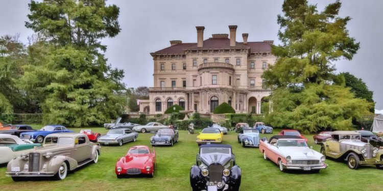 A variety of classic cars are displayed on a lawn in front of a large historic mansion on a cloudy day, capturing the spirit of East Coast Elegance: Audrain Newport Concours & Motor Week 2025.