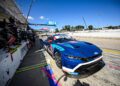 A blue Ford race car is stopped in the pit lane during Petit Le Mans, with crew members standing by and equipment ready on a sunny day.