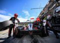 Race car pit stop at Petit Le Mans with crew members changing a rear tire and refueling the vehicle; Cadillac Racing and Rolex signage are visible in the background.