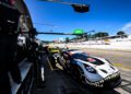 A black and white race car with a shark mouth design stops at the Petit Le Mans pit lane during a race, with crew members and another car in the background under a clear blue sky.