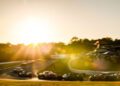 Sports cars race on a sunlit track at sunset during Petit Le Mans, with bright sunlight and lens flare affecting visibility.