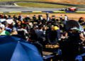 A race car speeds around the track at Petit Le Mans while a crowd of spectators, shaded by umbrellas, watches from the foreground on a sunny day.
