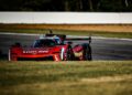 A red Whelen Cadillac racing car speeds along the Petit Le Mans track bordered by grass, with blurred trees, a white barrier, and the excitement of the Rolex event in the background.