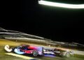 Two race cars drive side by side on the Petit Le Mans track at night, with long exposure light trails emphasizing their speed.