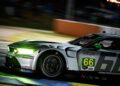 A silver race car with the number 66 speeds on the track at night during Petit Le Mans, its headlights cutting through the darkness and sponsor decals, including Rolex, visible on the side.