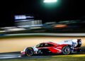 A Porsche racing car speeds along the track at night during Petit Le Mans, with blurred lights and surroundings emphasizing the high-speed motion.