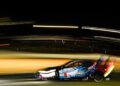 A race car with iHeartRadio branding speeds on the track at night during Petit Le Mans, headlights on, creating motion blur and light trails.